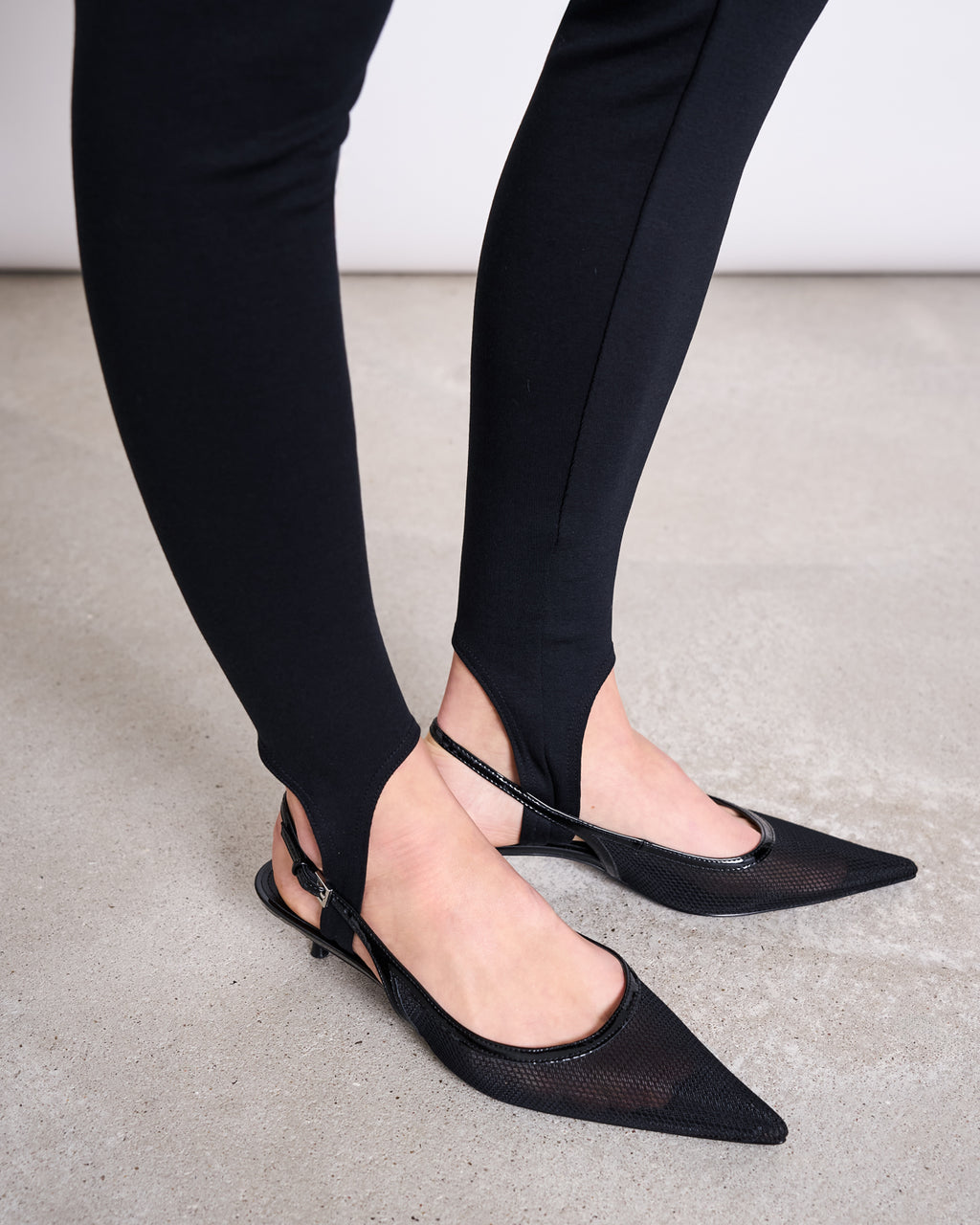 Someone wearing jan-n-june STIRRUP LEGGINGS BLACK, crafted from soft TENCEL™ Lyocell, and pointed-toe black slingback flats stands on a light gray concrete floor.