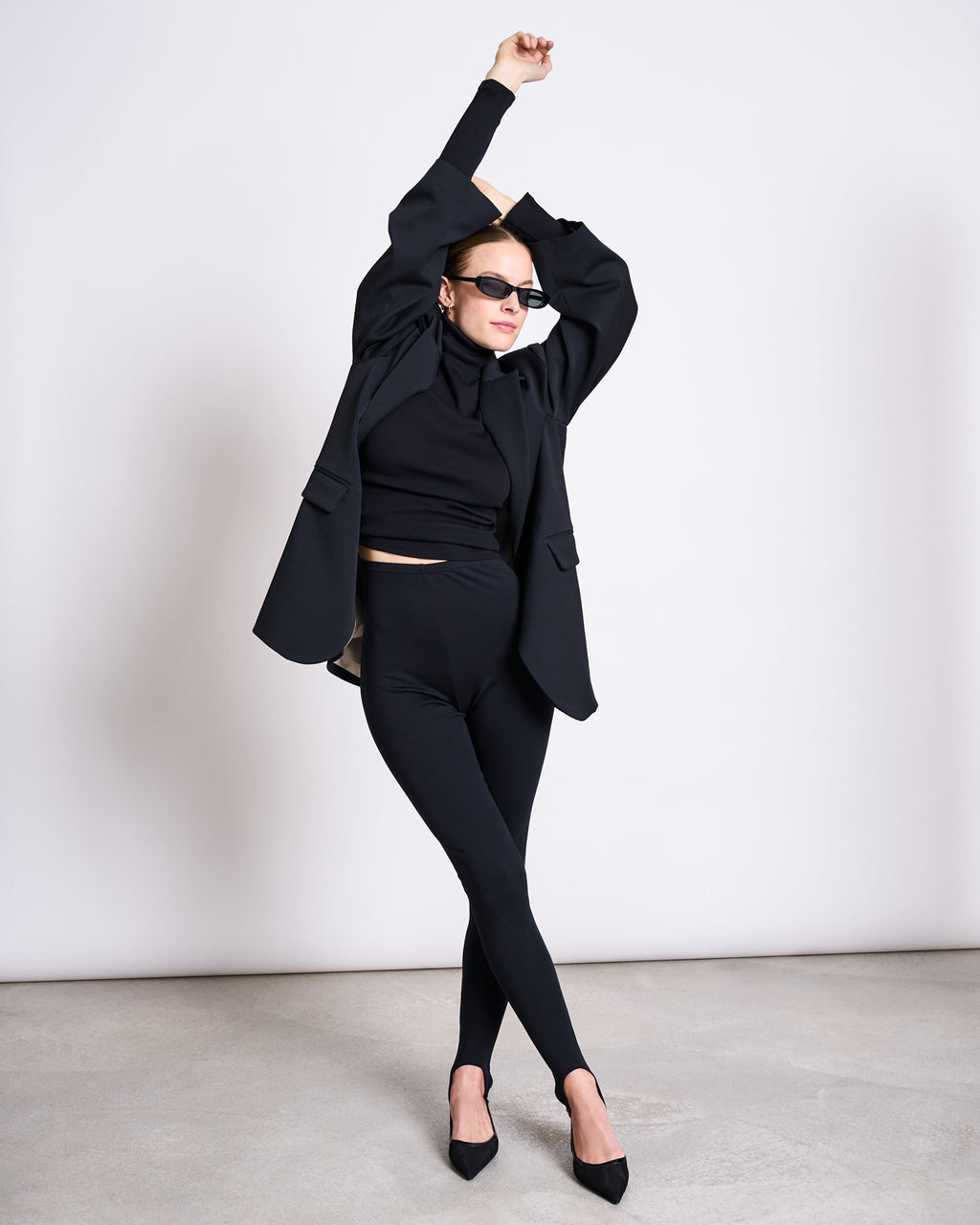 A woman poses against a white wall in jan-n-junes STIRRUP LEGGINGS BLACK, paired with a black blazer, turtleneck, black pointed heels, and sunglasses. She raises one arm overhead for a chic look.
