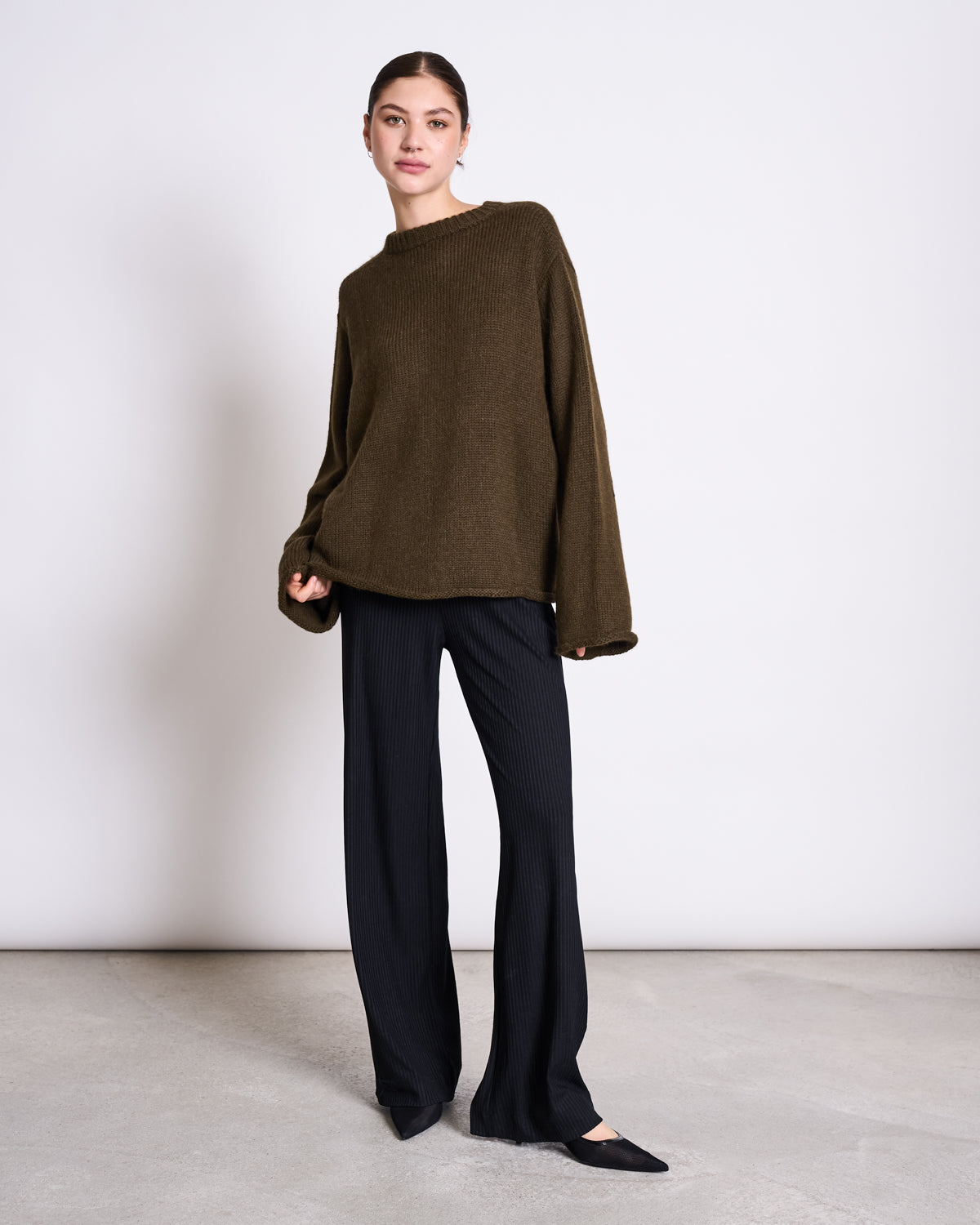 A woman poses in a minimalist room wearing the jan-n-june OVERSIZED ALPACA MIX JUMPER WOOLY SONA ROSEMARY, paired with black wide-leg pants and pointed shoes, her knee bent and head slightly tilted for a stylish look.
