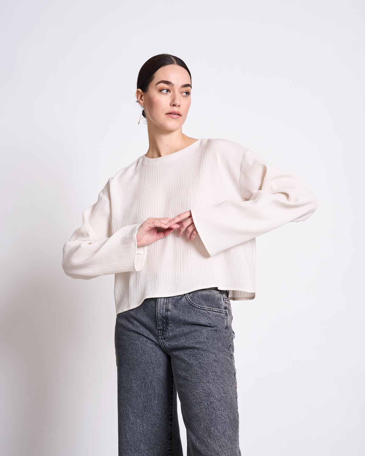 A woman with dark hair pulled back wears the jan-n-june CRINKLE BLOUSE YAME OFFWHITE, a loose, long-sleeved top, paired with high-waisted gray jeans. She stands against a white background, looking to the side with arms slightly raised.