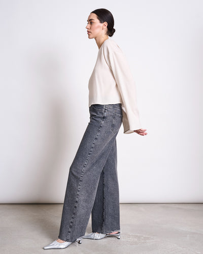 A woman with dark hair in a low bun poses sideways against a plain background, wearing the jan-n-june CRINKLE BLOUSE YAME OFFWHITE with wide-leg gray jeans and silver pointed heels. Her arms are relaxed behind her back.