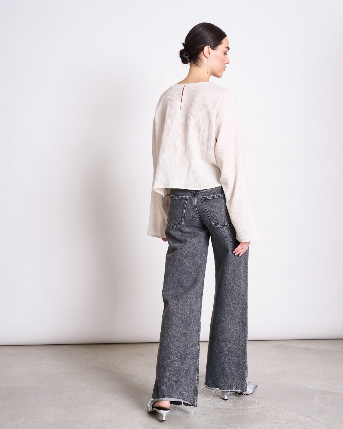 A woman faces away from the camera, wearing the jan-n-june CRINKLE BLOUSE YAME OFFWHITE with loose gray jeans and silver heels. Her hair is in a neat bun as she stands on a light floor against a plain wall.