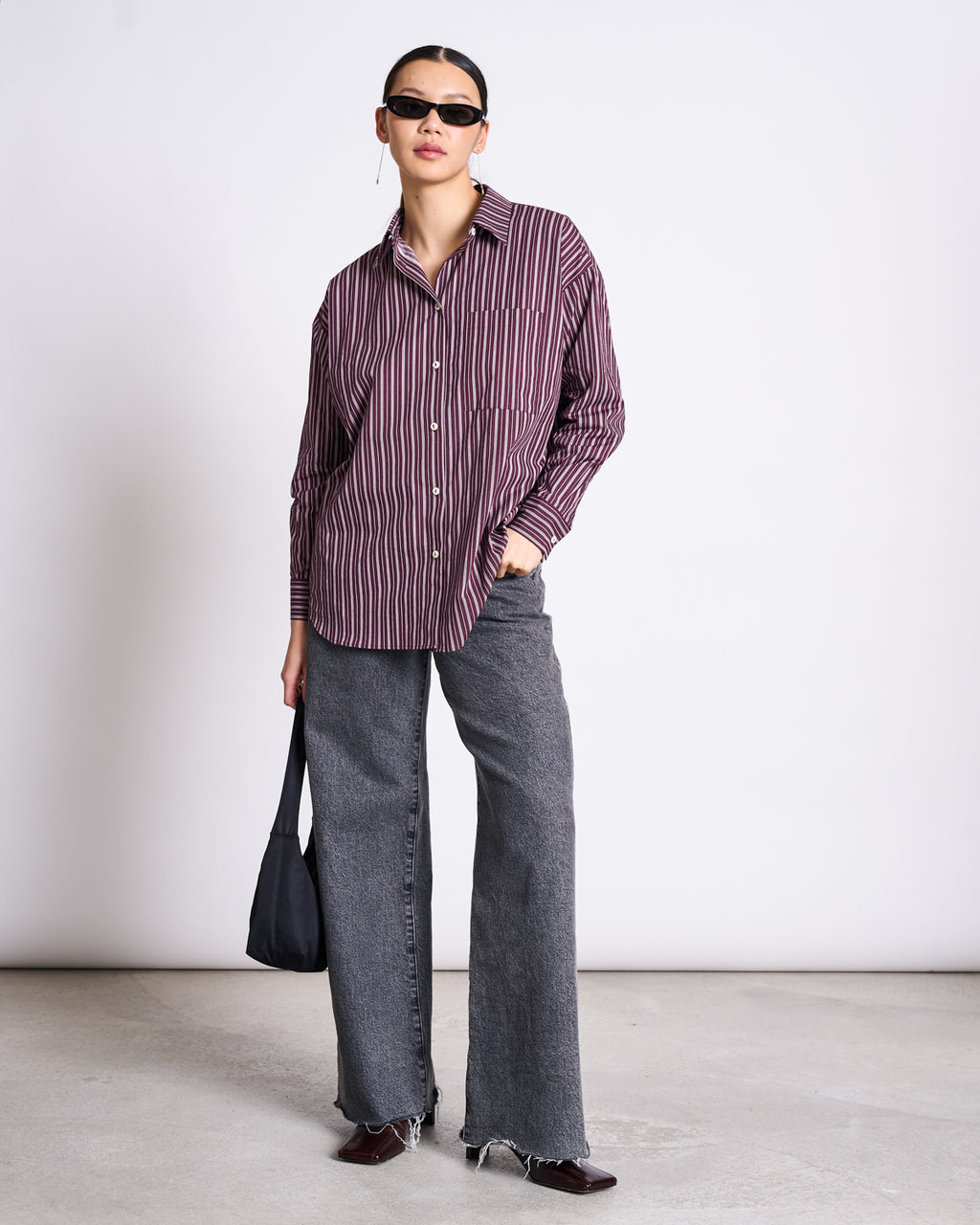 A person wearing black sunglasses, gray wide-leg jeans, dark shoes, and holding a black bag stands against a plain background in the jan-n-june OVERSIZED SHIRT MAMRO CHERRY LACQUER WHITE STRIPED.