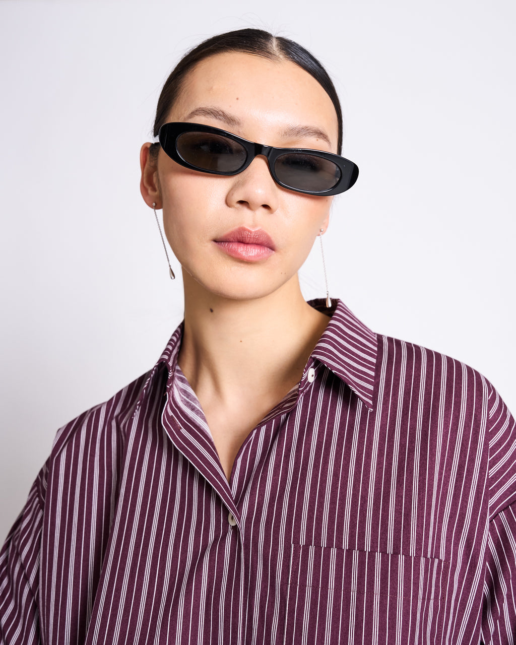A woman in black oval sunglasses, dangling earrings, and the jan-n-june OVERSIZED SHIRT MAMRO CHERRY LACQUER WHITE STRIPED poses against a plain white background.