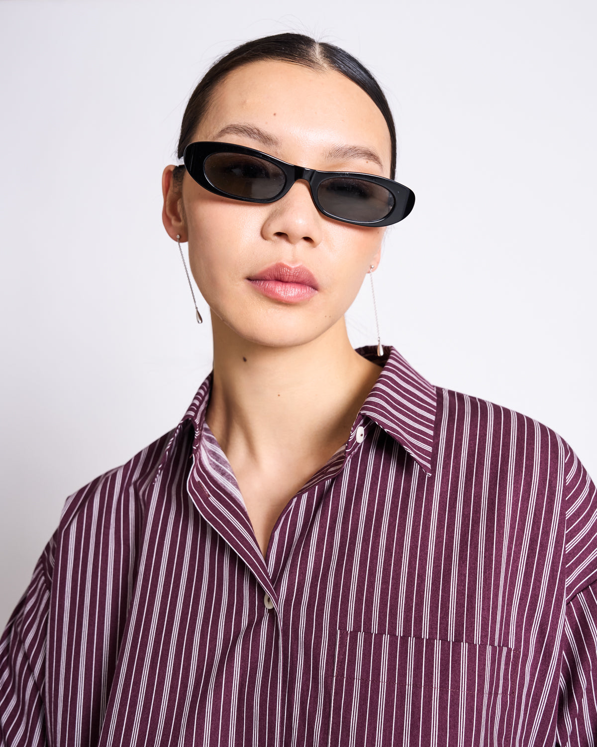 A woman in black oval sunglasses, dangling earrings, and the jan-n-june OVERSIZED SHIRT MAMRO CHERRY LACQUER WHITE STRIPED poses against a plain white background.