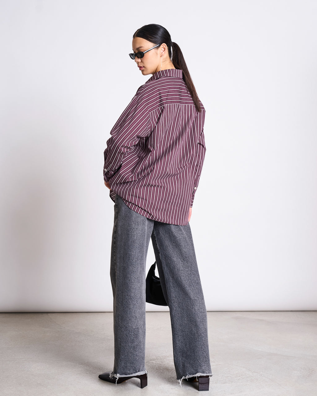 A woman stands sideways, looking back while wearing sunglasses, an OVERSIZED SHIRT MAMRO CHERRY LACQUER WHITE STRIPED by jan-n-june, wide-leg gray jeans with frayed hems, black heeled shoes, and holding a black handbag.