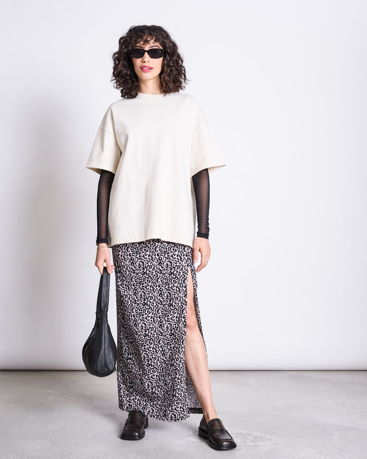 A woman with curly hair wears sunglasses, a loose cream T-shirt over a sheer black long-sleeve top, the jan-n-june MAXI SKIRT LUSTA LEO GOTS with a high slit, black loafers, and holds a black bag against a plain white background.