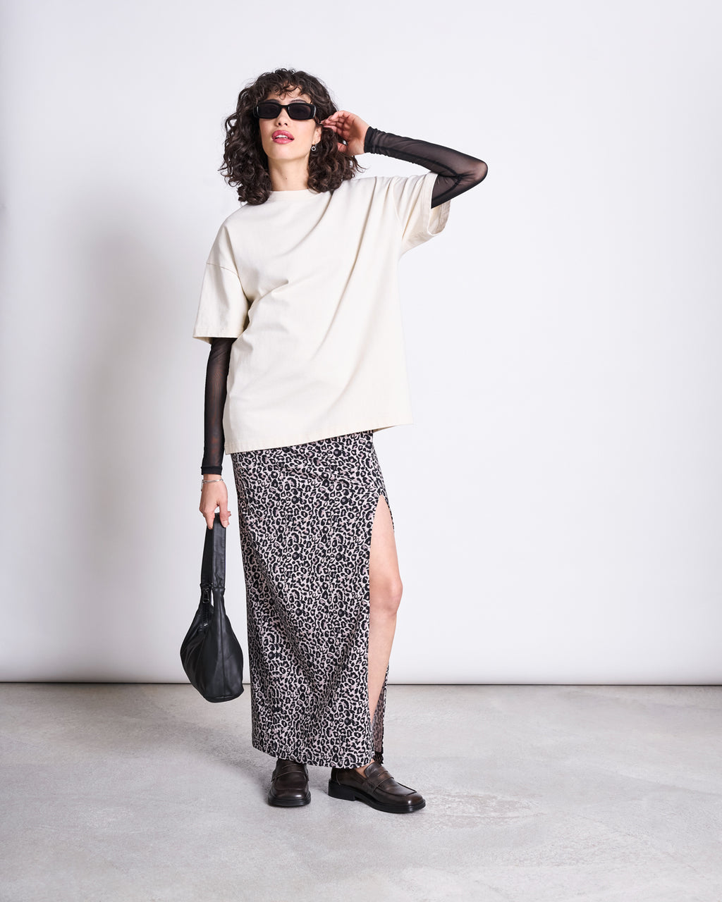 A woman wearing sunglasses, a loose white t-shirt over a black mesh long-sleeve, the jan-n-june MAXI SKIRT LUSTA LEO GOTS, black shoes, and holding a black bag stands against a plain white background.