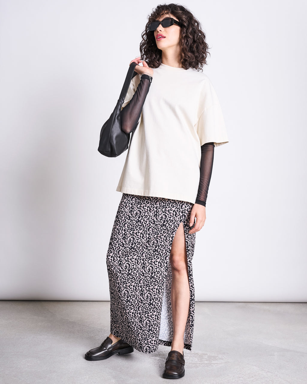 A woman in sunglasses wears a cream oversized tee, sheer black sleeves, and the jan-n-june MAXI SKIRT LUSTA LEO GOTS—a high-waisted leo print maxi with a side slit—paired with black loafers and a black shoulder bag against a plain background.