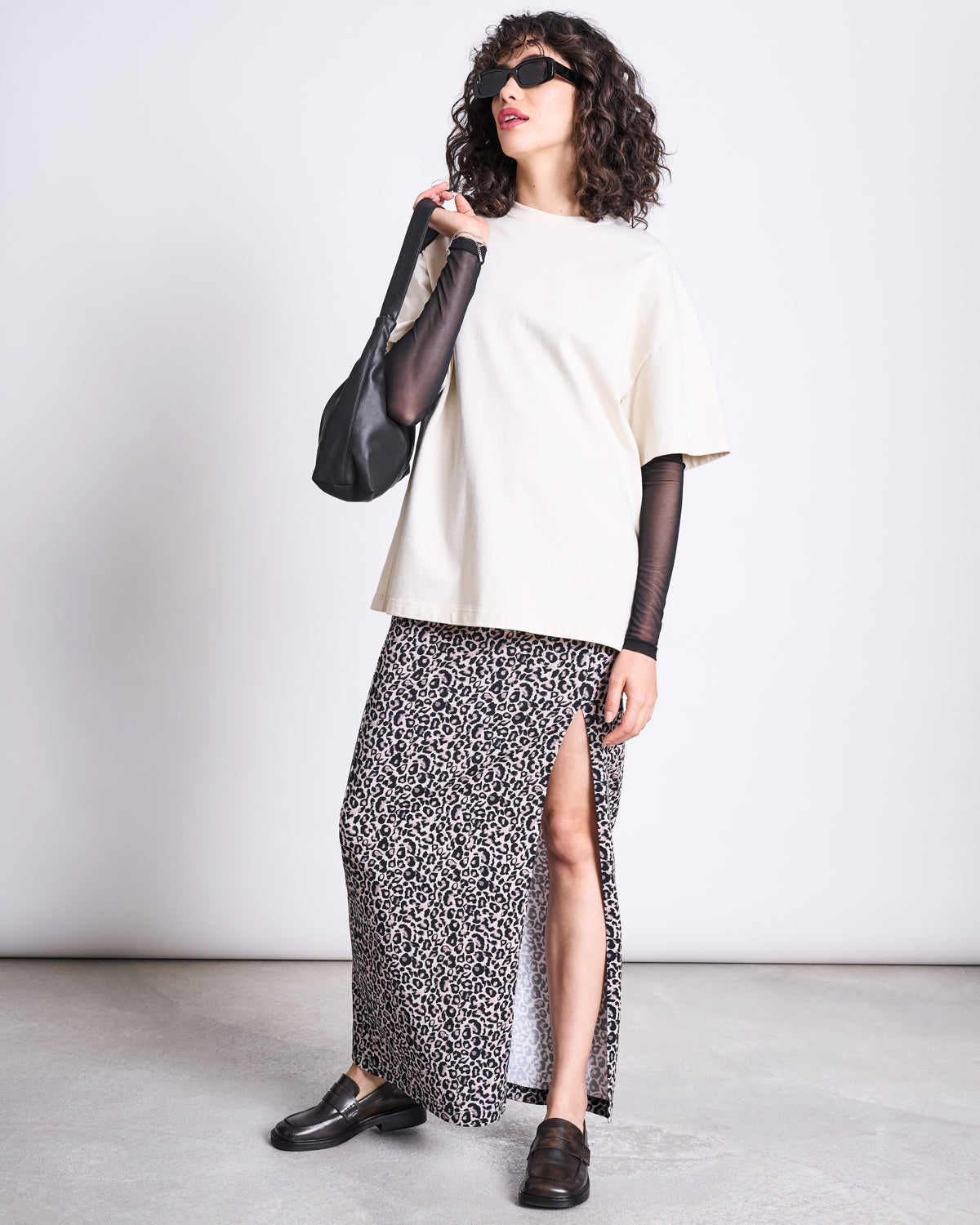 A woman in sunglasses wears a cream oversized tee, sheer black sleeves, and the jan-n-june MAXI SKIRT LUSTA LEO GOTS—a high-waisted leo print maxi with a side slit—paired with black loafers and a black shoulder bag against a plain background.