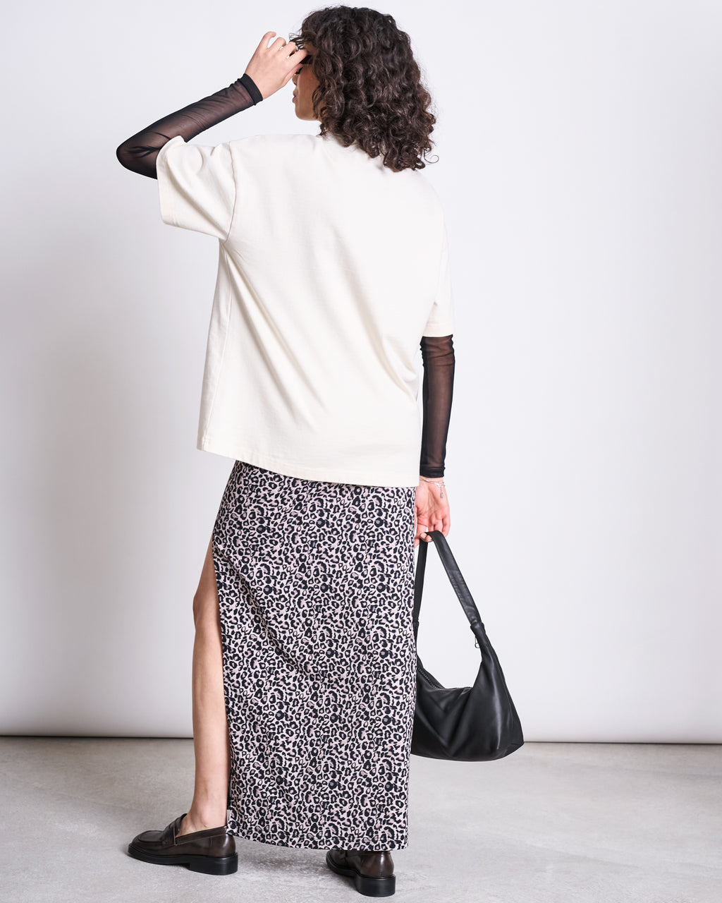 A person faces away, wearing a loose white tee over sheer black sleeves, the jan-n-june MAXI SKIRT LUSTA LEO GOTS—a high-waisted leo print maxi skirt with side slit—paired with black loafers and a black shoulder bag.
