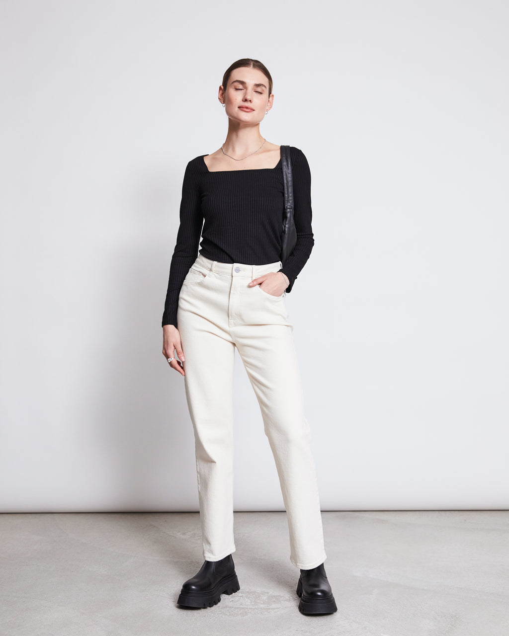 A person stands against a white background wearing the jan-n-june SQUARE NECK LONGSLEEVE FRIA BLACK top, off-white high-waisted jeans, and black platform boots. One hand is in a pocket while the other rests by their side.