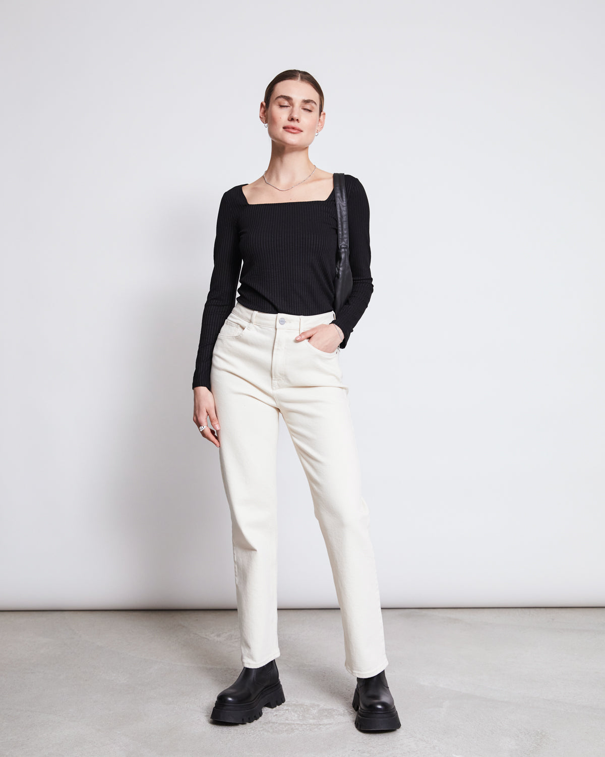 A person stands against a white background wearing the jan-n-june SQUARE NECK LONGSLEEVE FRIA BLACK top, off-white high-waisted jeans, and black platform boots. One hand is in a pocket while the other rests by their side.