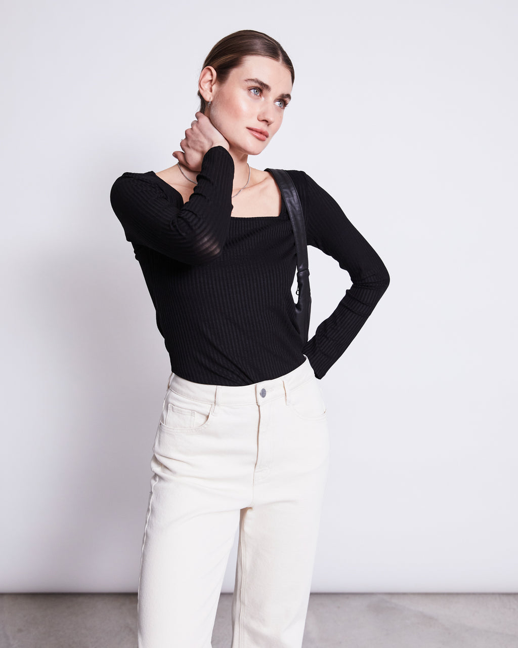 A person wears the jan-n-june SQUARE NECK LONGSLEEVE FRIA BLACK with high-waisted white jeans, standing against a plain background, looking to the side and touching their neck while carrying a black shoulder bag.