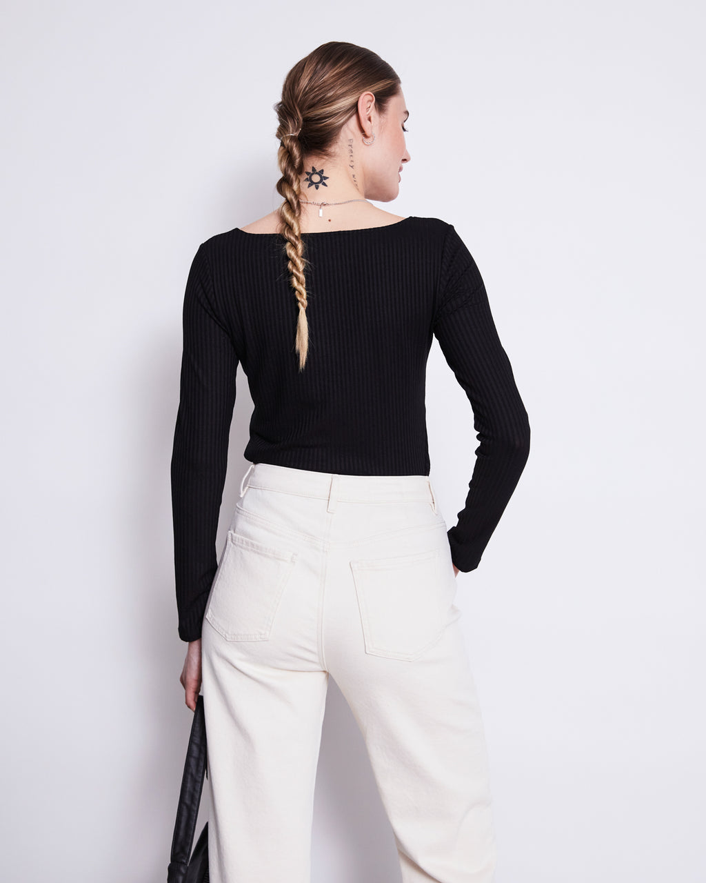 A woman with a braided ponytail and sun tattoo on her neck models the jan-n-june SQUARE NECK LONGSLEEVE FRIA BLACK top with high-waisted white pants, standing with her back to the camera against a plain white background.
