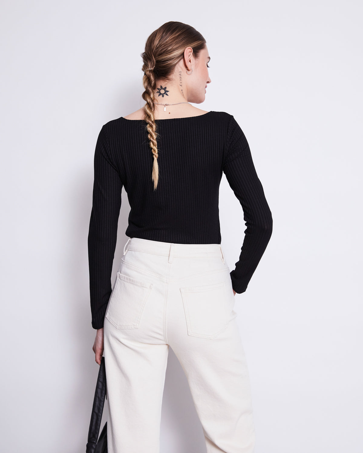 A woman with a braided ponytail and sun tattoo on her neck models the jan-n-june SQUARE NECK LONGSLEEVE FRIA BLACK top with high-waisted white pants, standing with her back to the camera against a plain white background.