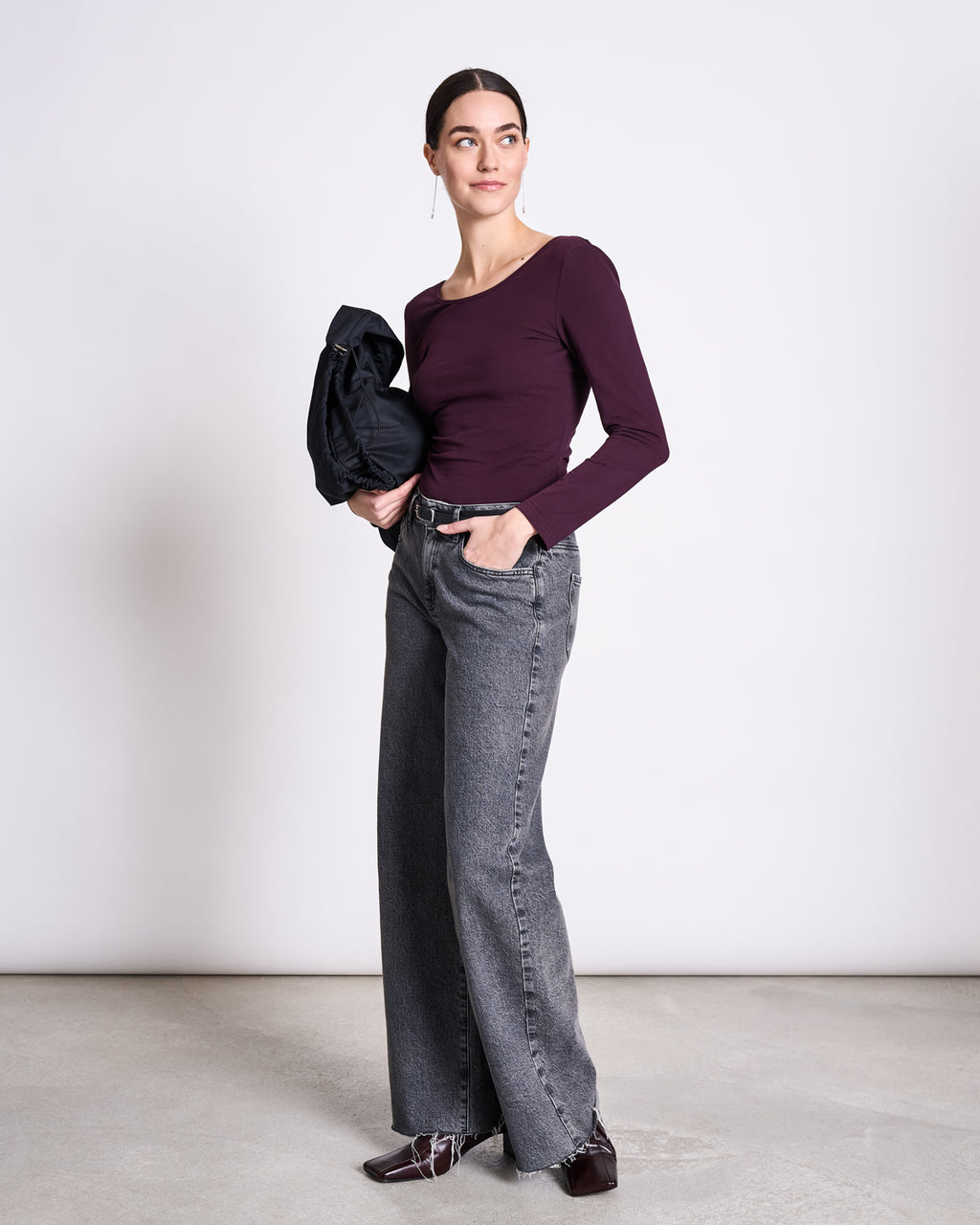 A woman stands against a plain background, wearing the jan-n-june BALLERINA LONGSLEEVE LEON in cherry lacquer, gray wide-leg jeans, and brown shoes, holding a dark jacket over her left arm with her right hand in her pocket.