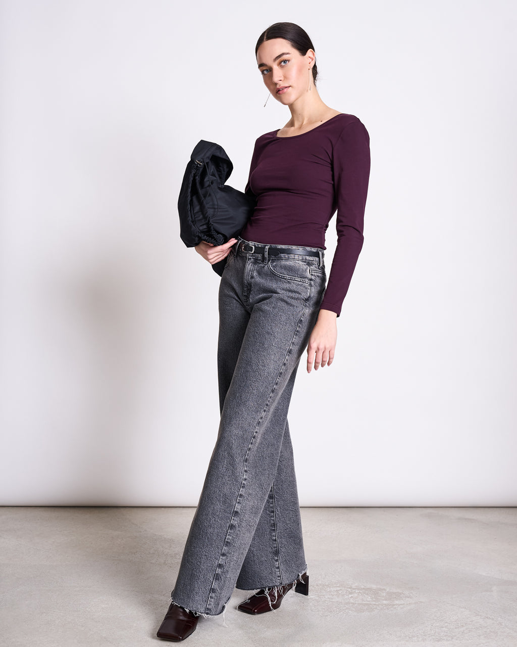 A woman stands against a plain background, confidently wearing the jan-n-june BALLERINA LONGSLEEVE LEON CHERRY LACQUER GOTS with gray high-waisted wide-leg jeans, dark shoes, and holding a black jacket.