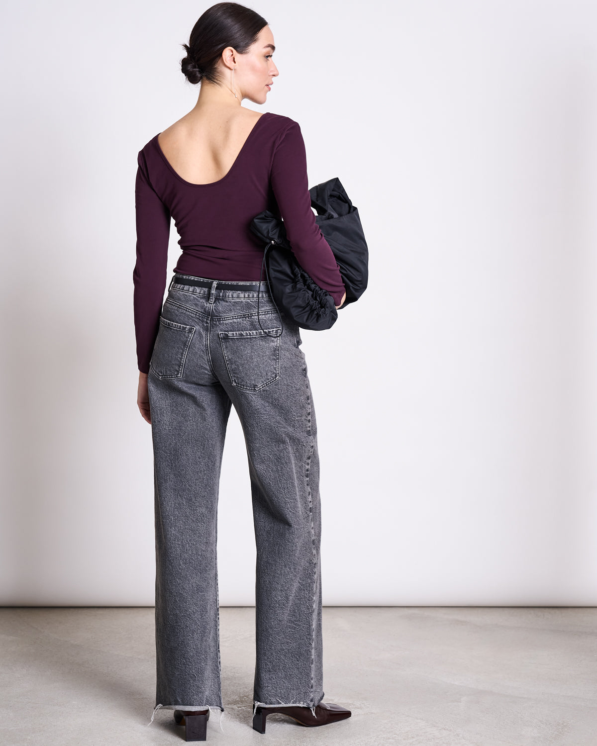 A woman faces away, wearing the jan-n-june BALLERINA LONGSLEEVE LEON CHERRY LACQUER GOTS in organic cotton, gray high-waisted jeans, and brown pointed boots. She holds a black bag, with her hair in a low bun against a neutral background.