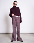 A person in dark sunglasses and a maroon chunky knit sweater poses casually with one hand in their pocket, wearing jan-n-june’s COTTON PANTS SOBY CHERRY LACQUER WHITE against a plain light background.
