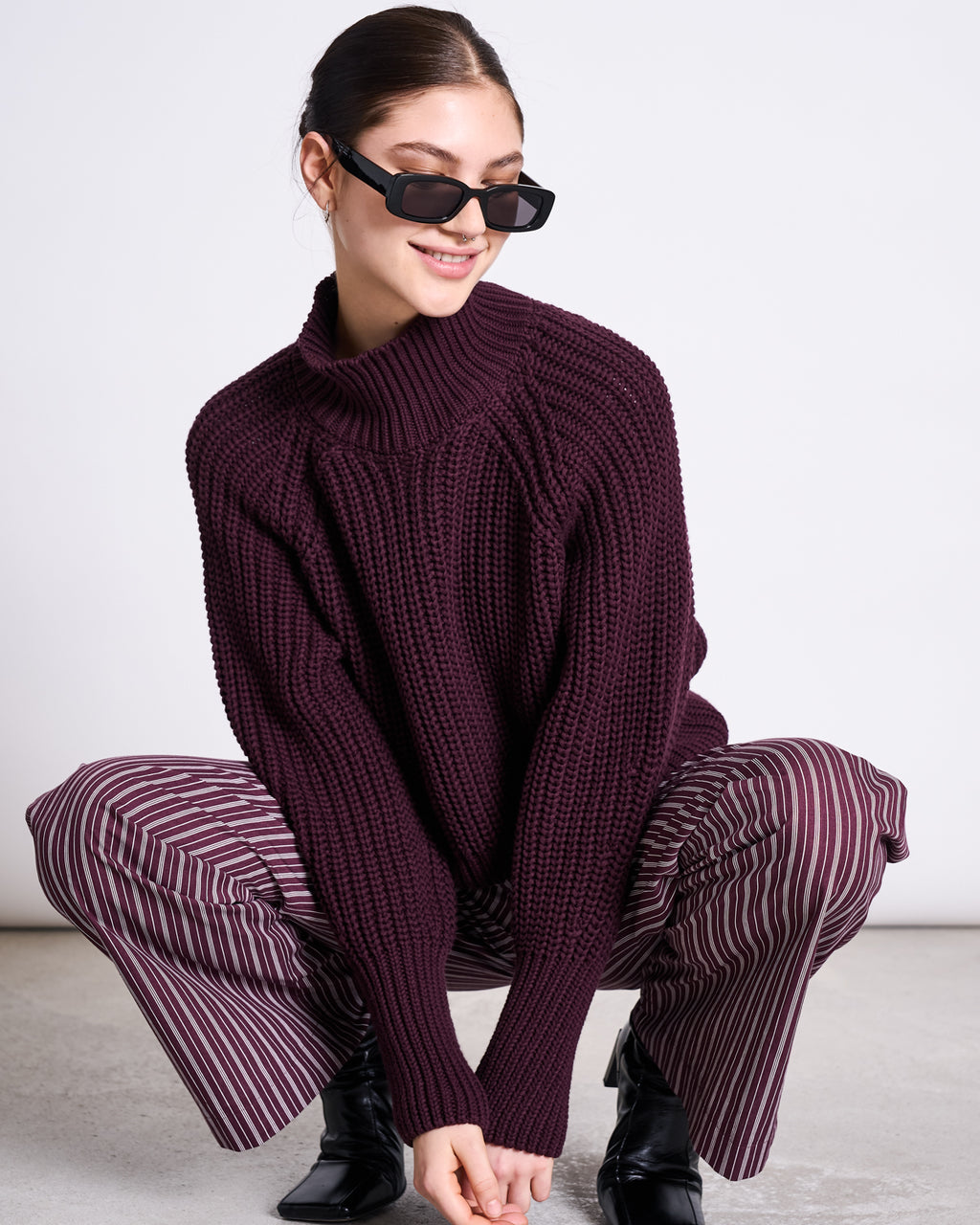 A woman in black sunglasses and a dark purple chunky knit sweater squats and smiles against a white background, wearing jan-n-june’s COTTON PANTS SOBY CHERRY LACQUER WHITE.