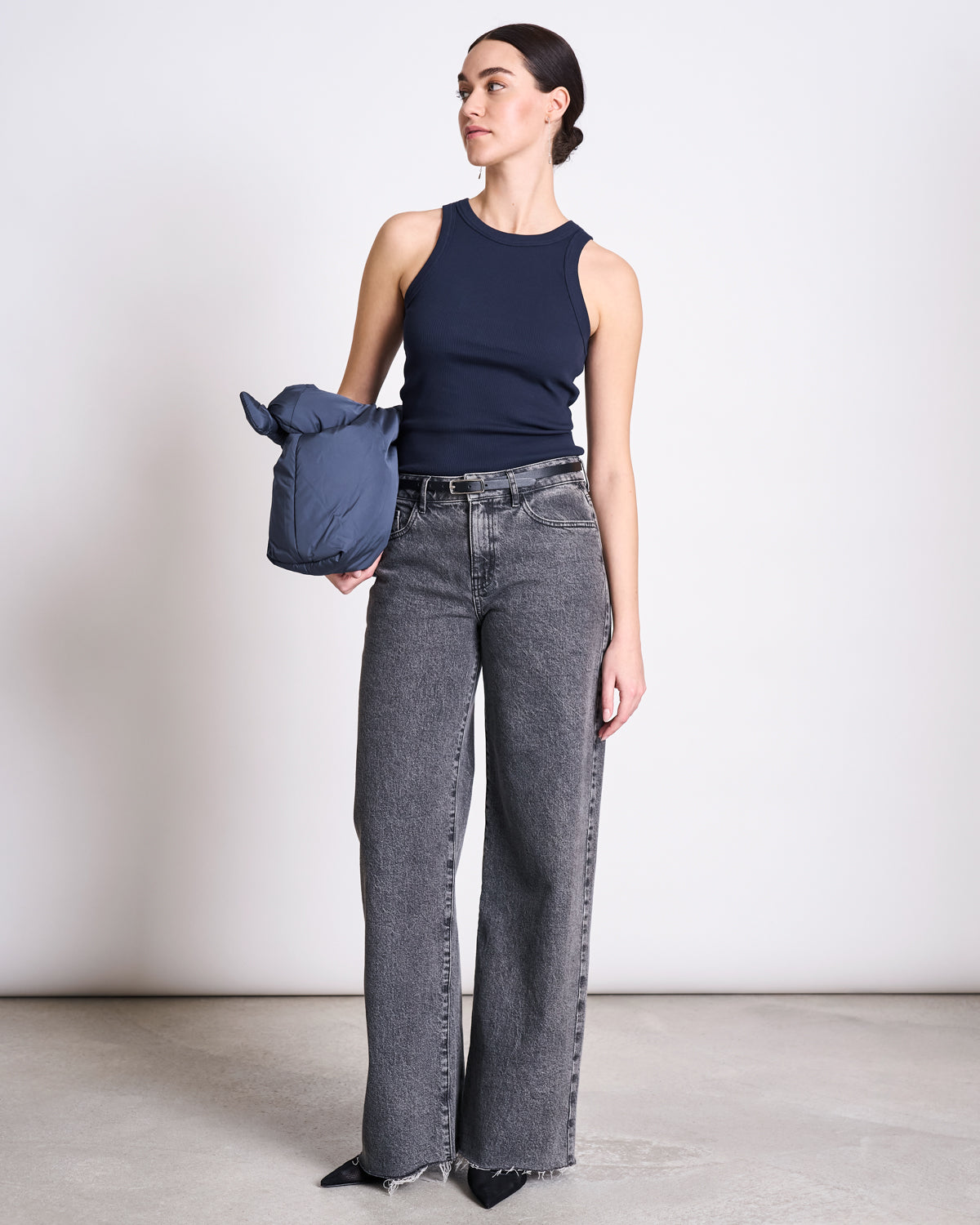 A woman stands against a plain background wearing the jan-n-june ORGANIC COTTON TANK TOP PORTO LONG NIGHTFALL GOTS, high-waisted wide-leg gray jeans, holding a blue jacket and wearing black pointed-toe shoes.