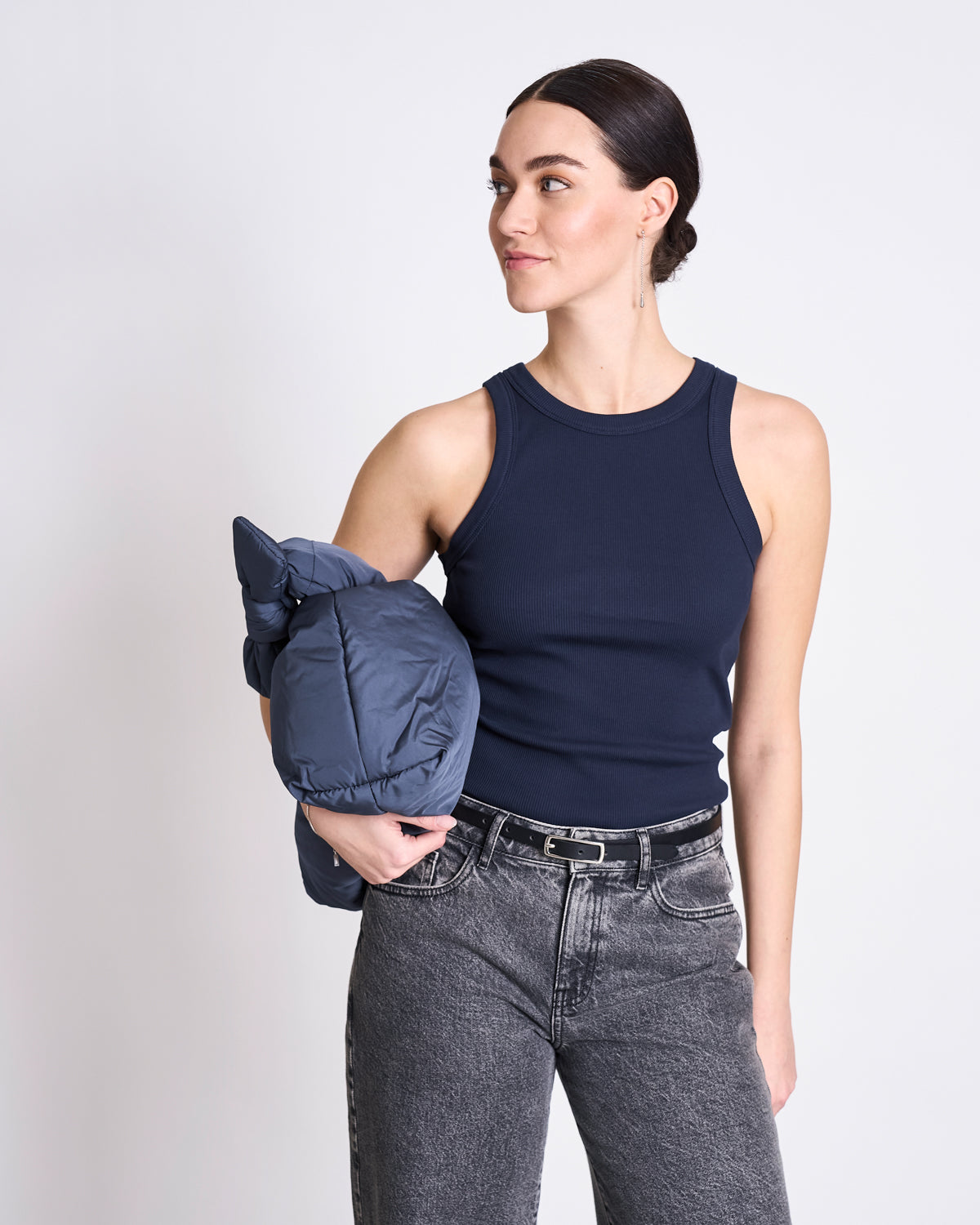 A woman with dark hair tied back wears the jan-n-june ORGANIC COTTON TANK TOP PORTO LONG NIGHTFALL GOTS in navy, paired with gray jeans. She holds a blue puffer jacket, standing against a plain light background and looking right.