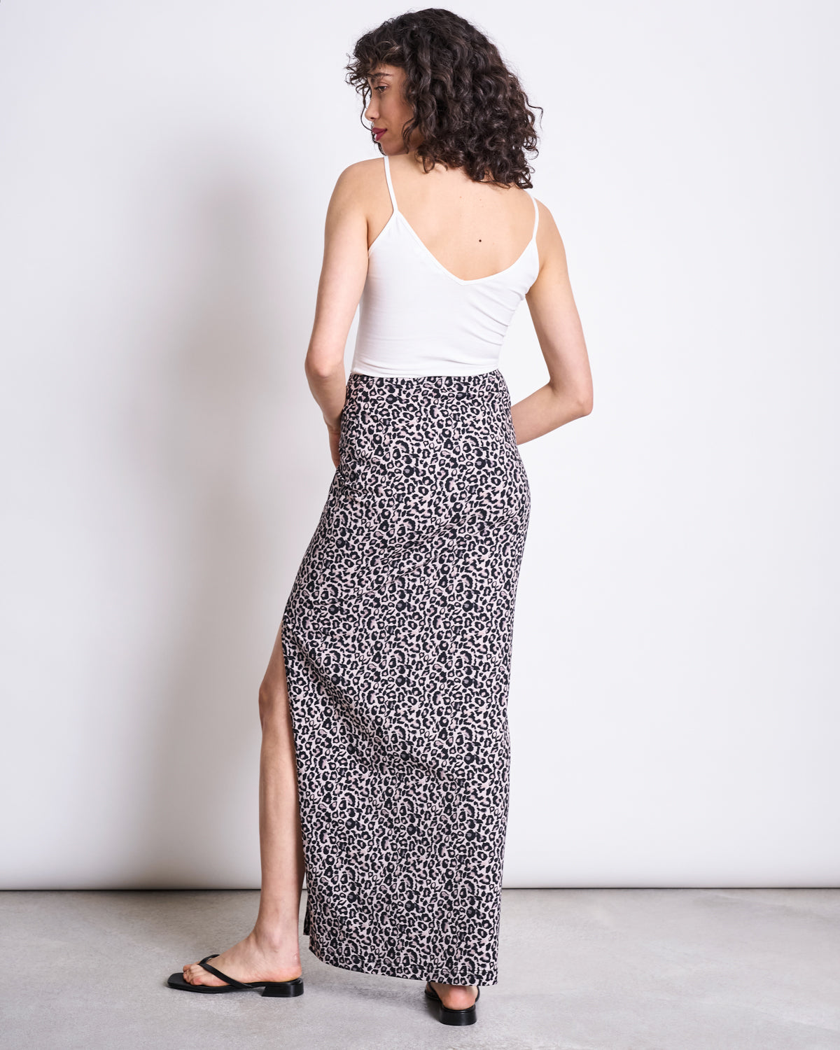 A woman faces away, wearing a white sleeveless top and the jan-n-june MAXI SKIRT LUSTA LEO GOTS—a high-waisted, leopard print maxi skirt with a side slit—paired with black sandals against a light background.