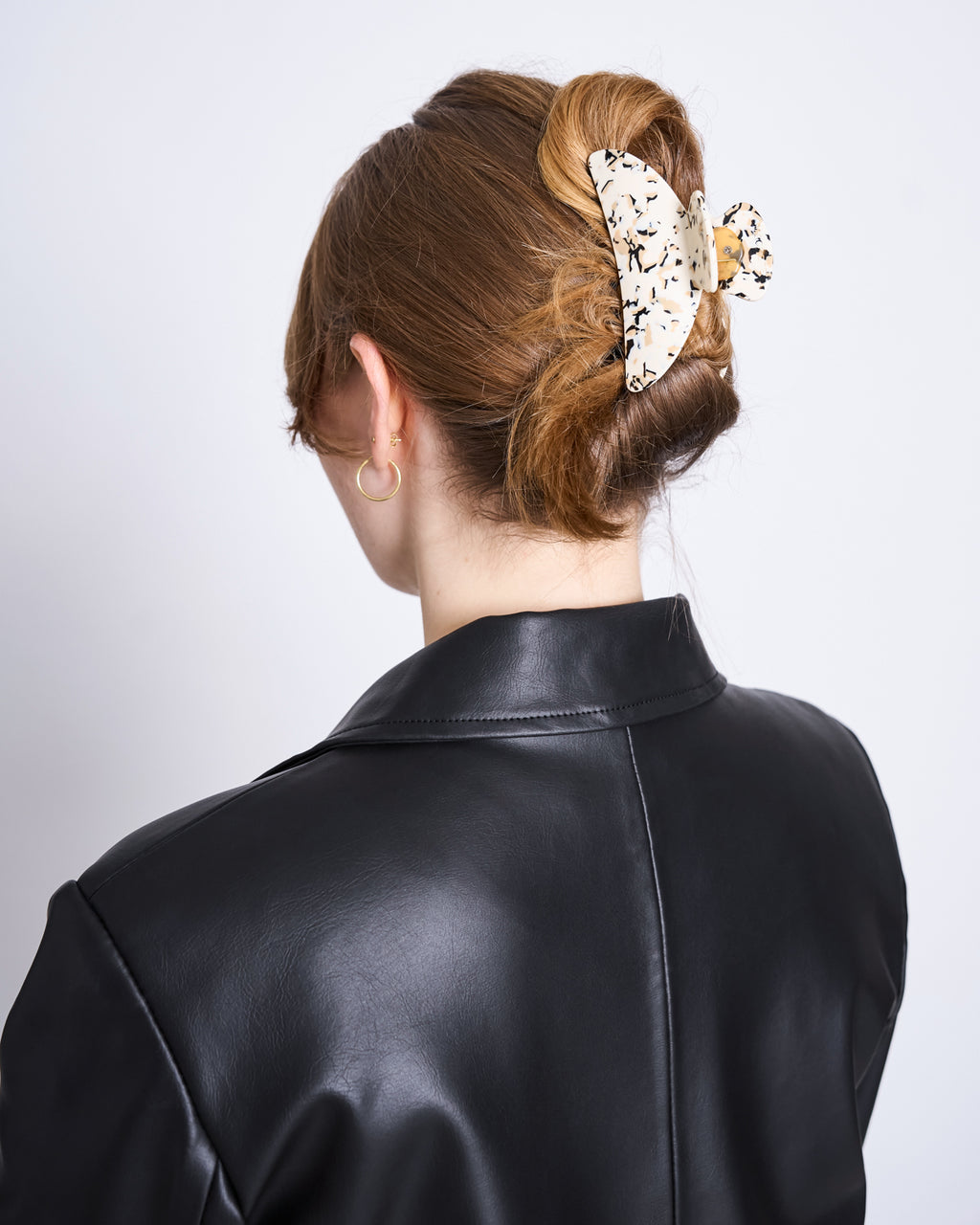 A person with light brown hair in a low updo, secured by the elegant jan-n-june MIDI HEIRLOOM CLAW TERRAZZO. Seen from behind, they wear a black leather jacket and gold hoop earrings against a plain background.