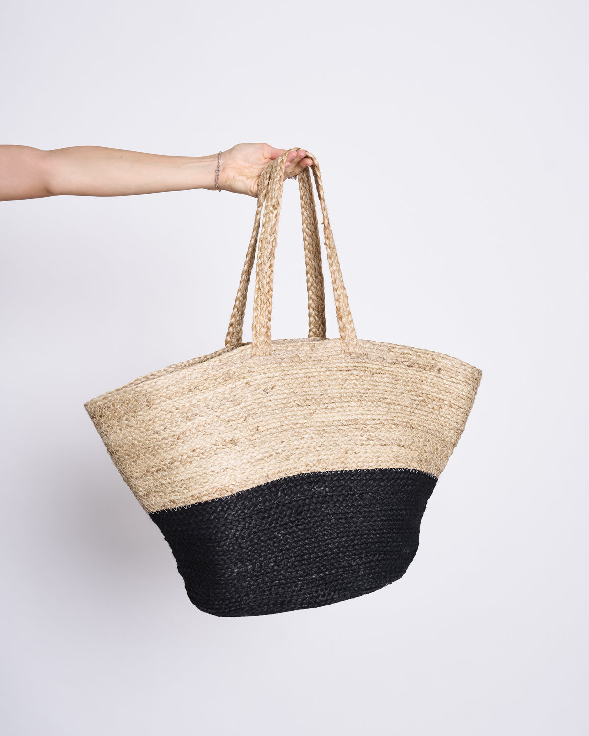 A hand holds the jan-n-june SHOPPING BASKET JUTE BLACK, featuring two handles and a design with a natural straw top and black bottom. Made under a Women’s Empowerment programme, it is set against a plain white background.