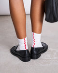Close-up of a person wearing jan-n-june OVERLOCK SOCKS WHITE CHERRY RED—organic cotton socks with red wavy trim—styled with black loafers and a black bag, standing on a light-colored floor.