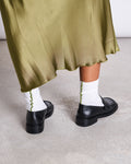 A person stands on a gray concrete floor, wearing shiny olive green skirt, black loafers, and jan-n-june OVERLOCK SOCKS WHITE WOODY OLIVE—comfy organic cotton socks with green wavy trim.