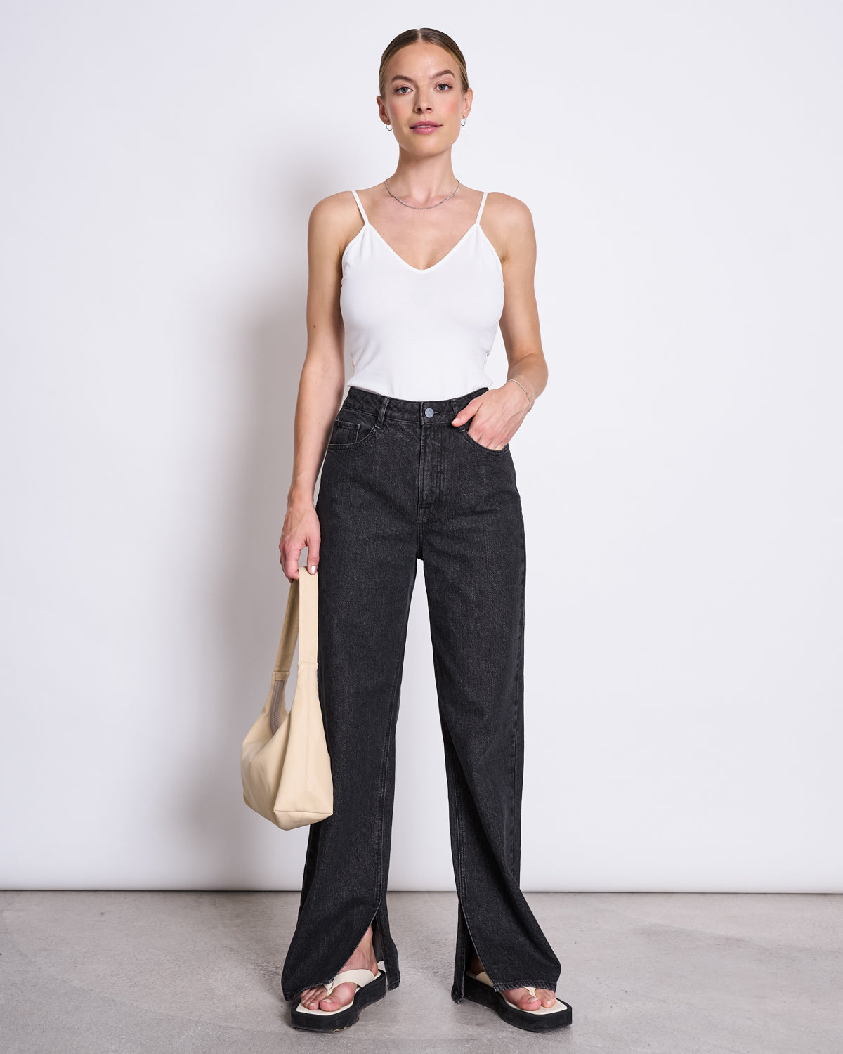 A woman stands against a white wall, wearing the jan-n-june TOP MATILDA WHITE GOTS V-neck jersey camisole made from organic cotton. She pairs it with high-waisted black jeans, black sandals, and a beige shoulder bag, her hair tied back as she looks at the camera.