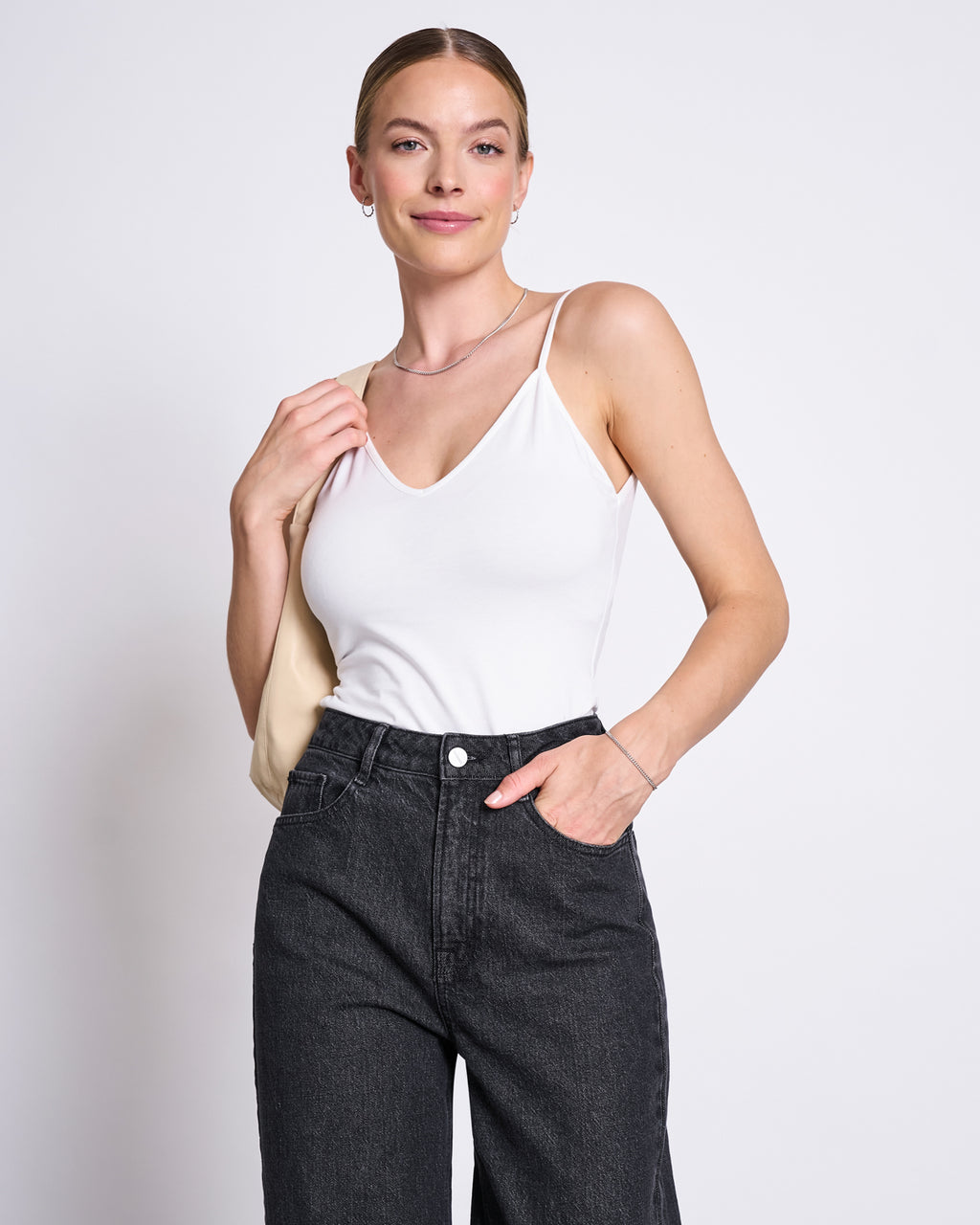 A woman with light skin and blonde hair in a low bun smiles in front of a plain light background, wearing jan-n-junes TOP MATILDA WHITE GOTS with high-waisted black jeans and holding a beige bag over her shoulder.