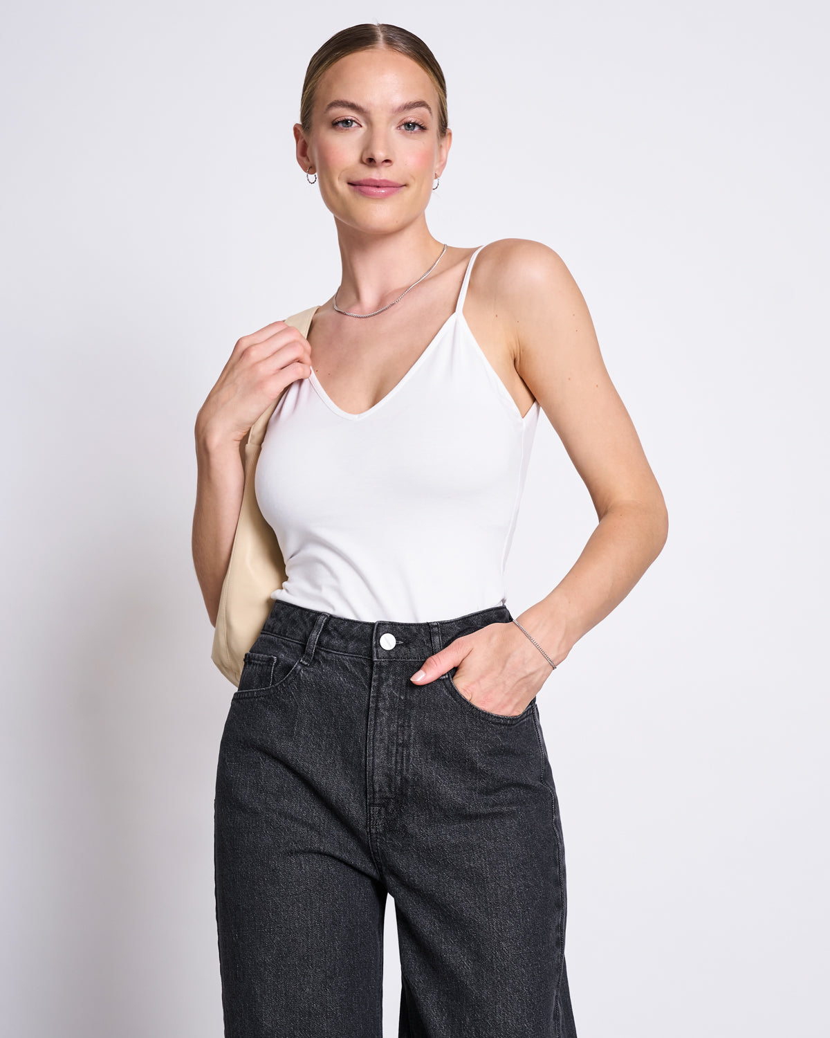 A woman with light skin and blonde hair in a low bun smiles in front of a plain light background, wearing jan-n-junes TOP MATILDA WHITE GOTS with high-waisted black jeans and holding a beige bag over her shoulder.