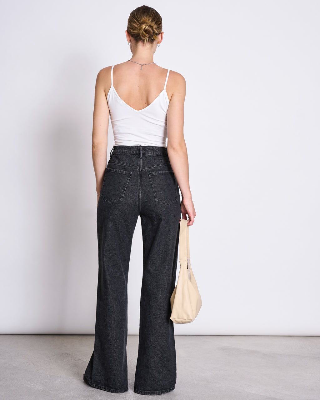A woman with her hair in a bun faces away from the camera, wearing the jan-n-june TOP MATILDA WHITE GOTS V-neck jersey and high-waisted black jeans, holding a light handbag in her left hand.