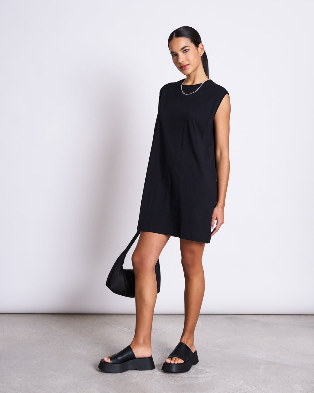 A woman with straight dark hair pulled back stands confidently against a white background, wearing the jan-n-june MINI DRESS NANITI BLACK, black platform sandals, a silver necklace, and holding a black bag.