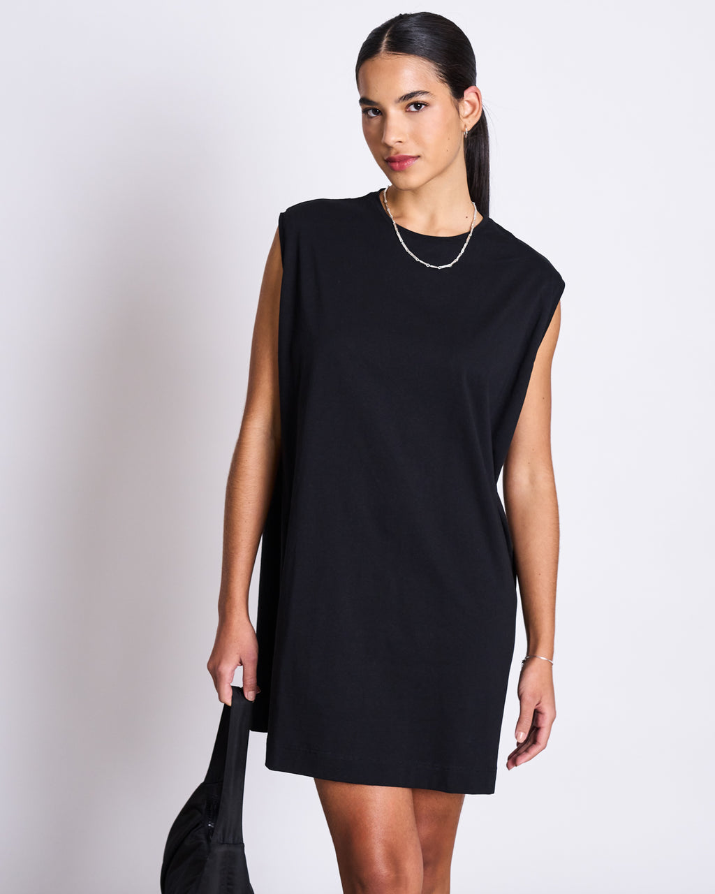 A woman with dark hair wears the jan-n-june MINI DRESS NANITI BLACK and a simple gold necklace, holding a black bag. She stands against a white background, looking confidently at the camera—perfect summer style.