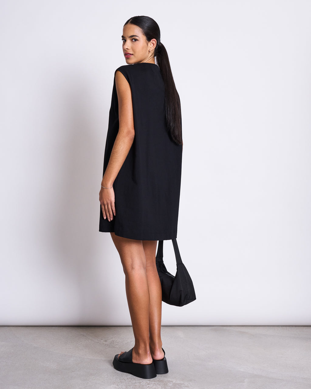 A woman with long dark hair tied back faces away, glancing over her shoulder. She wears the jan-n-june MINI DRESS NANITI BLACK with platform shoes and a small black handbag. A plain light background highlights her effortless summer style.