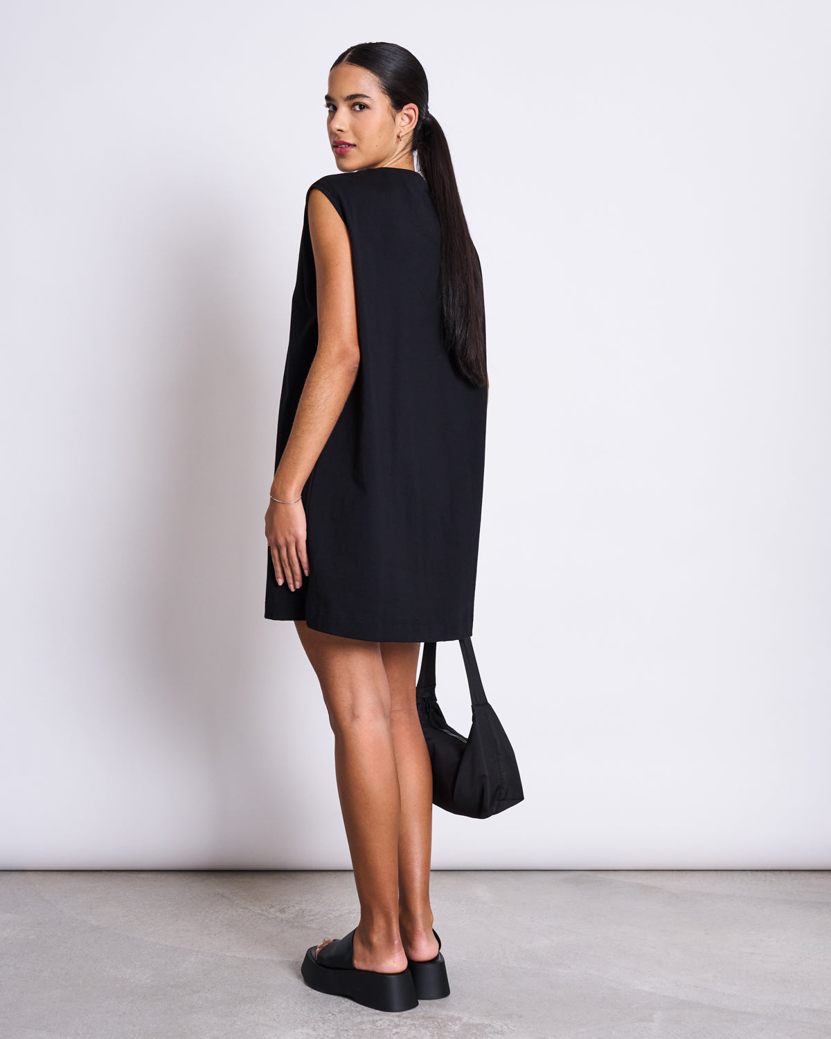 A woman with long dark hair tied back faces away, glancing over her shoulder. She wears the jan-n-june MINI DRESS NANITI BLACK with platform shoes and a small black handbag. A plain light background highlights her effortless summer style.