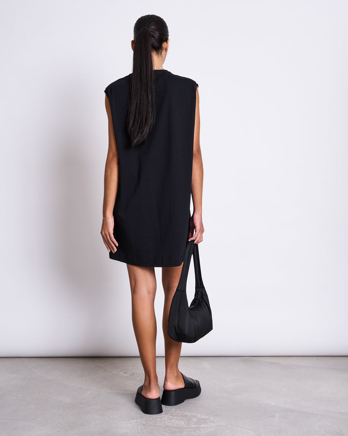 A woman with long dark hair in a ponytail stands facing away, wearing the jan-n-june MINI DRESS NANITI BLACK—ideal for summer—paired with black slides and a small black handbag, against a plain white wall and light floor.