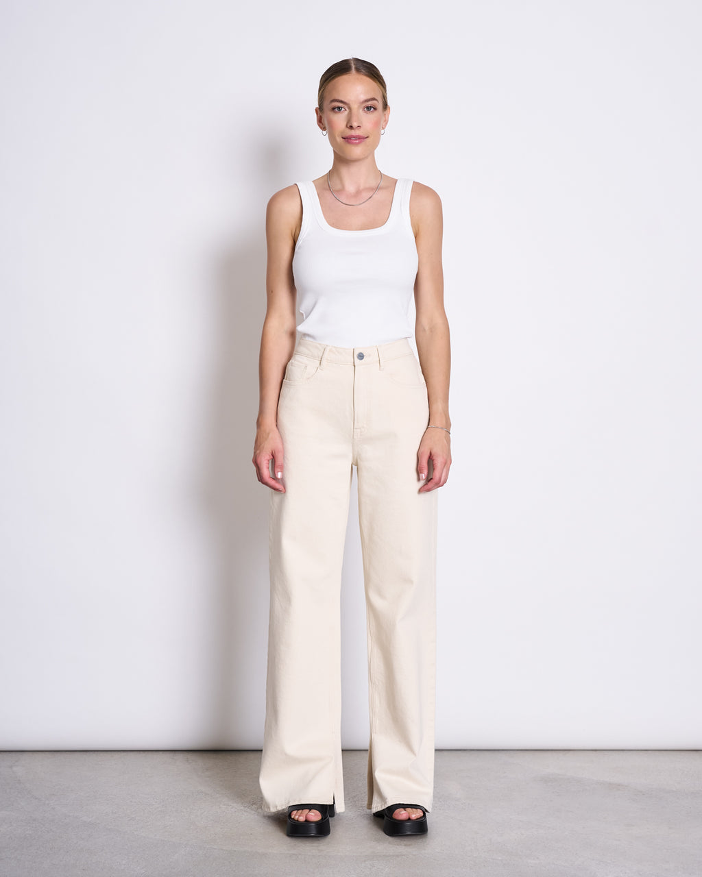 A woman stands against a white wall, wearing the jan-n-june SLITTED WIDE LEG JEANS SELENE ECRU and a white tank top with black platform sandals. She looks straight ahead, arms relaxed at her sides.