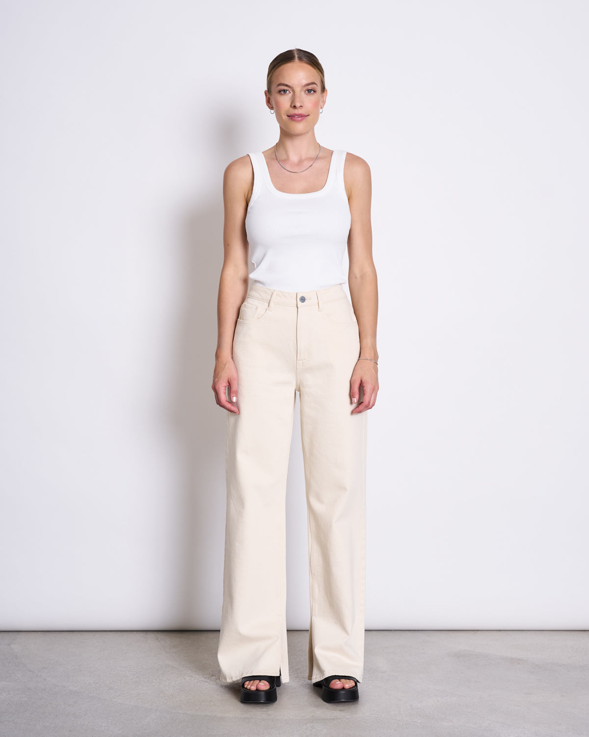A woman stands against a white wall, wearing the jan-n-june SLITTED WIDE LEG JEANS SELENE ECRU and a white tank top with black platform sandals. She looks straight ahead, arms relaxed at her sides.