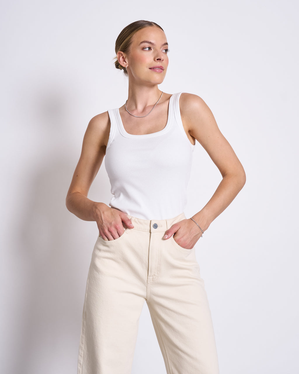 A woman stands against a plain white background, wearing the jan-n-june SLITTED WIDE LEG JEANS SELENE ECRU and a white sleeveless tank top. She looks to the side with a slight smile, her hands in her pockets.