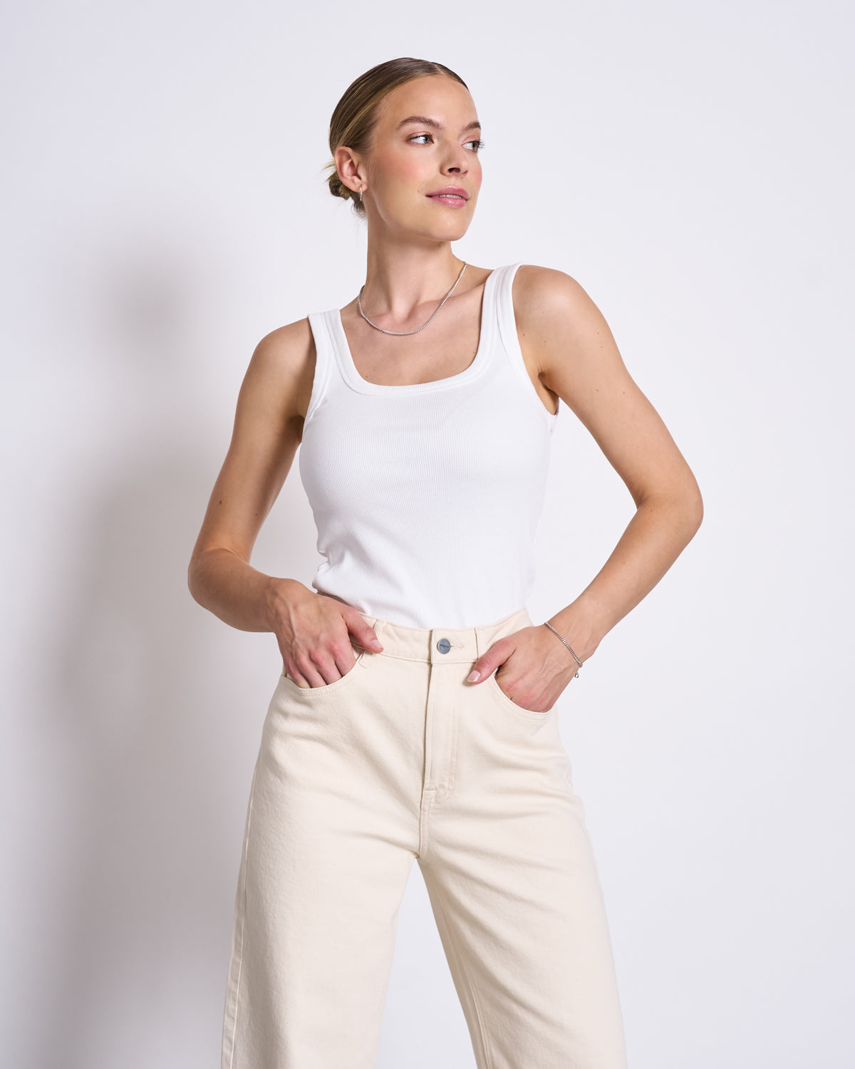 A woman stands against a plain white background, wearing the jan-n-june SLITTED WIDE LEG JEANS SELENE ECRU and a white sleeveless tank top. She looks to the side with a slight smile, her hands in her pockets.