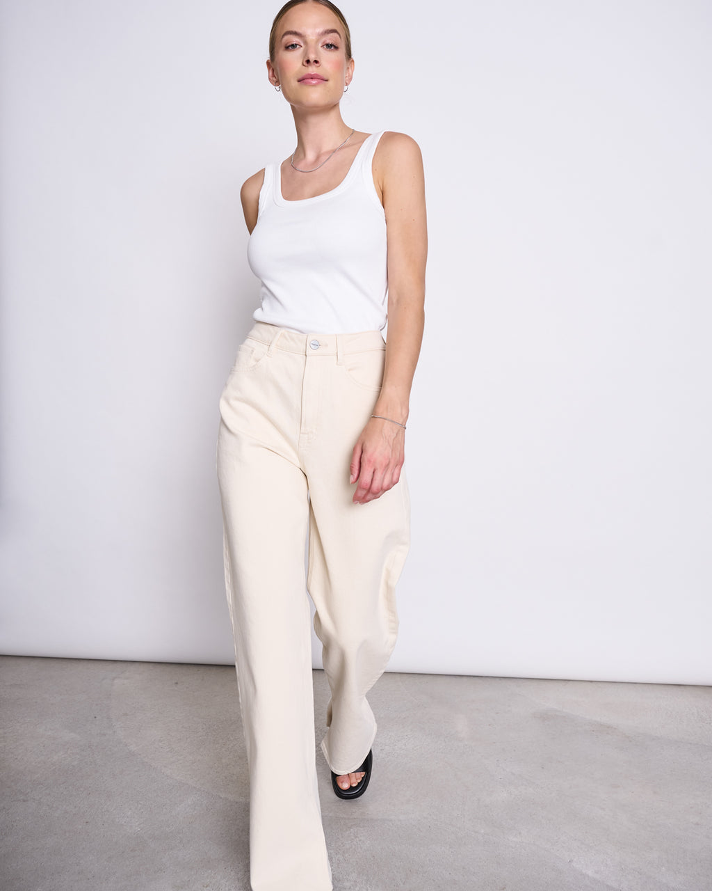 A woman stands against a white background, wearing jan-n-junes SLITTED WIDE LEG JEANS SELENE ECRU with a white sleeveless tank top and black shoes. She looks forward with a relaxed expression, her hair tied back.