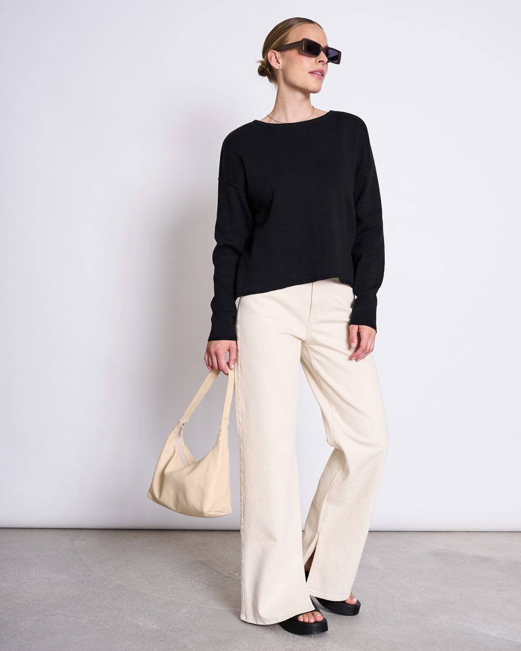 Dressed in jan-n-june’s LIGHT KNIT JUMPER WILLIAMSBURG BLACK GOTS, a woman with sunglasses and hair pulled back stands against a plain wall, styled with cream wide-leg pants, black sandals, and a cream handbag.