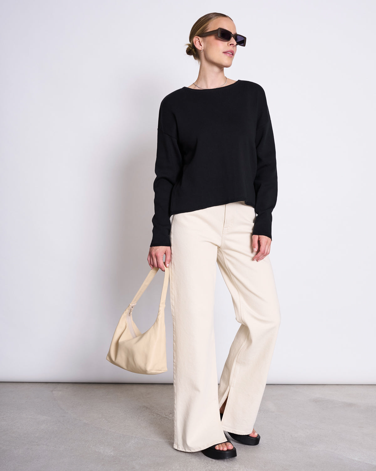 Dressed in jan-n-june’s LIGHT KNIT JUMPER WILLIAMSBURG BLACK GOTS, a woman with sunglasses and hair pulled back stands against a plain wall, styled with cream wide-leg pants, black sandals, and a cream handbag.