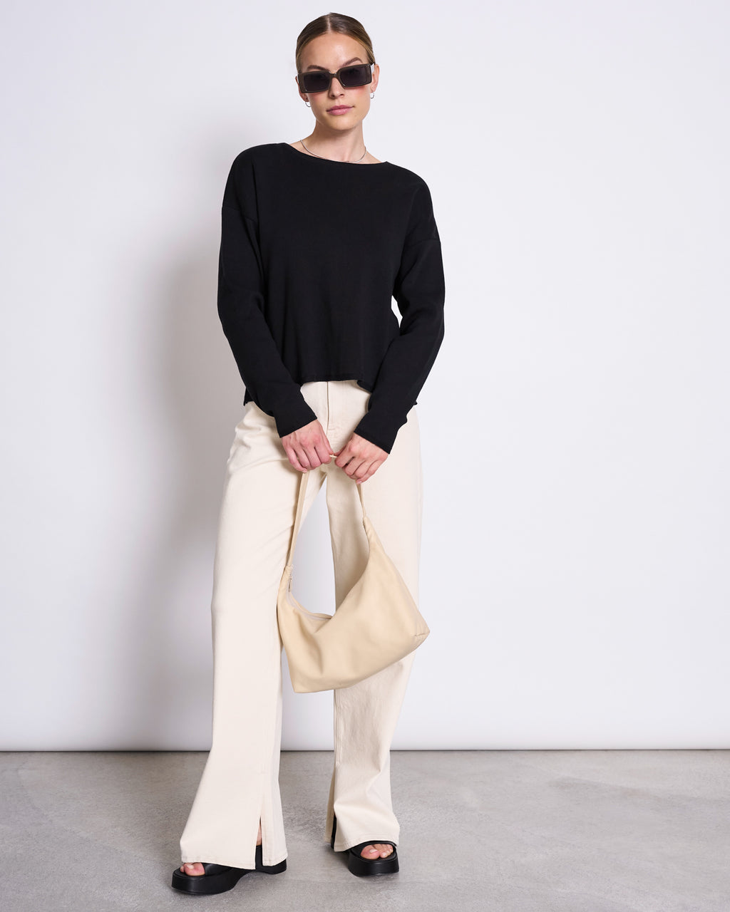 A woman stands against a plain wall wearing sunglasses, the jan-n-june LIGHT KNIT JUMPER WILLIAMSBURG BLACK GOTS, cream wide-leg pants with front slits, black platform sandals, and a beige handbag—an ideal capsule wardrobe look.