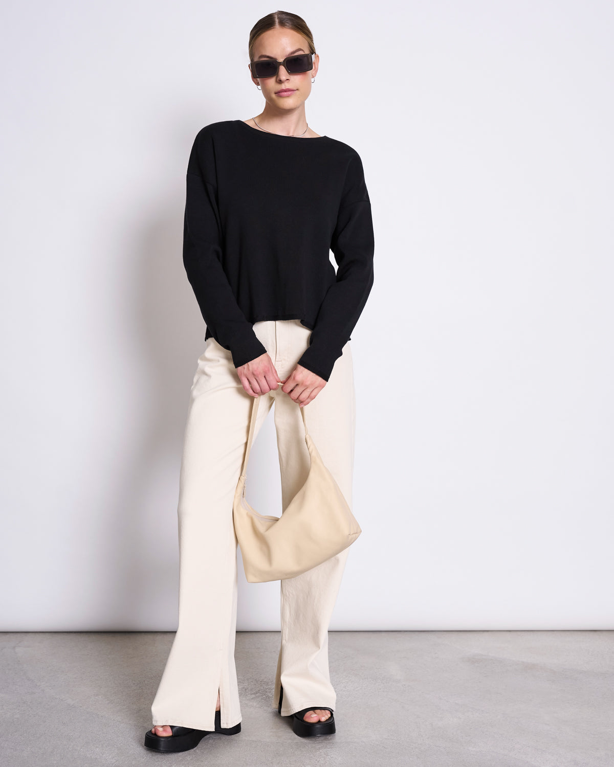 A woman stands against a plain wall wearing sunglasses, the jan-n-june LIGHT KNIT JUMPER WILLIAMSBURG BLACK GOTS, cream wide-leg pants with front slits, black platform sandals, and a beige handbag—an ideal capsule wardrobe look.